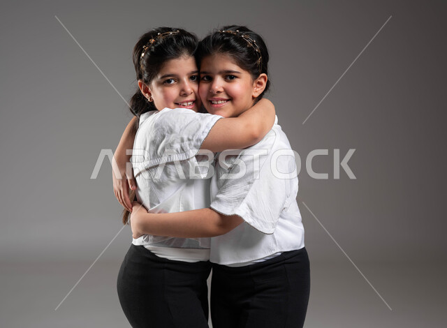 مفهوم الصداقة والأخوة، تقوية وتعزيز الروابط العائلية، بورتريه مقرب لتؤام بنات عربيات خليجيات سعوديات مبتسمات يقومون بمعانقة بعضهما بحرارة، النظر الى الكاميرا بتعابير المحبة والسرور، التعاطف والمودة بين الشقيقات، خلفية رمادية
