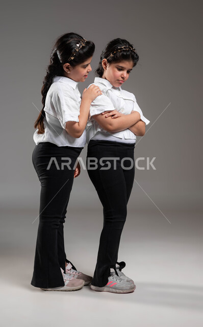 الخصومة وسوء التفاهم بين الشقيقات، محاولة الوصول الى تفاهم وصلح بين الاختان المتخاصمتان، بورتريه لفتاة عربية خليجية سعودية تقوم بمحاولة التحدث الى اختها التوأم، تعابير الحزن والزعل، حل المشاكل بطريقة فعالة، صورة الجسم بالطول الكامل، خلفية رمادية