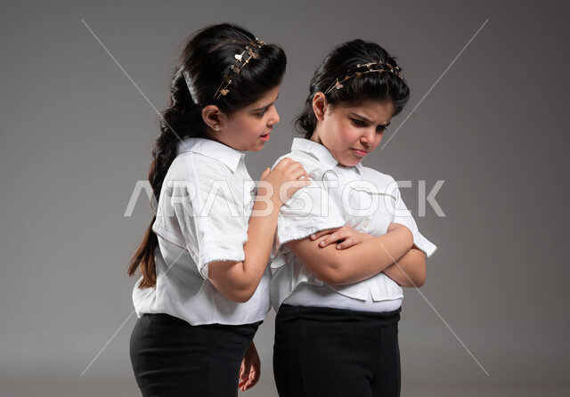 Solving problems in an effective way, rivalry and misunderstanding between sisters, trying to reach an understanding and reconciliation between two quarreling sisters, a close-up portrait of a Saudi Arabian Gulf girl trying to talk to her twin sister, expressions of sadness and anger, gray background.