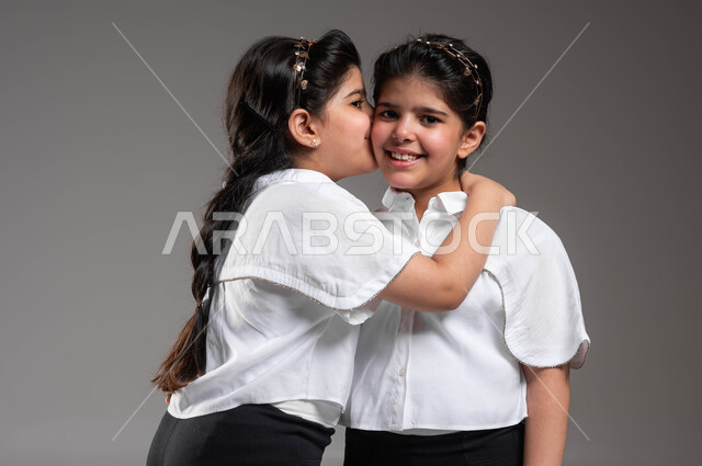 علاقة عائلية مبينية على الألفة والمودة، ايماءات تدل على العطف والحنان، مفهوم الترابط الأسري والمحبة بين الأخوة، بورتريه مقرب لفتاة عربية خليجية سعودية تقبل أختها التوأم بحرارة، النظر الى الكاميرا بتعابير السرور والسعادة، خلفية رمادية