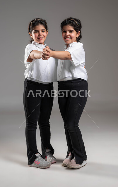Strengthening family ties through play and recreational activities, spending enjoyable times with sisters, the concept of true friendship between brothers, a portrait of two smiling Saudi Gulf Arab twin sisters holding each other’s hands looking at the camera with expressions of pleasure, full body portrait, gray background
