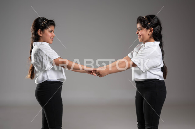 قضاء أوقات ممتعة مع الشقيقات، مفهوم الصداقة و الأخوة، تقوية الروابط العائلية من خلال اللعب والأنشطة الترفيهية، بورتريه مقرب لأختين توأم عربيتين خليجيتين سعوديتين تمسكان بأيدي بعضهما بمحبة، خلفية رمادية