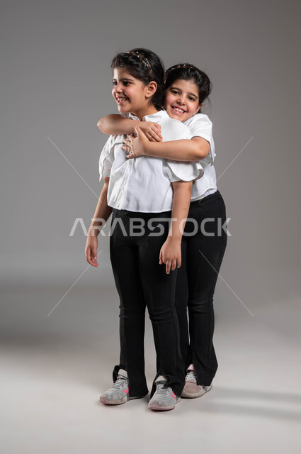 التعاطف والمودة بين الشقيقات، مفهوم الصداقة و الأخوة، تقوية وتعزيز الروابط العائلية، بورتريه لفتاة عربية خليجية سعودية مبتسمة تعانق اختها التوأم وتنظر اللى الكاميرا بتعابير المحبة والسرور، مفهوم مساندة ودعم الأخوات، صورة الجسم بالكامل، خلفية رمادية