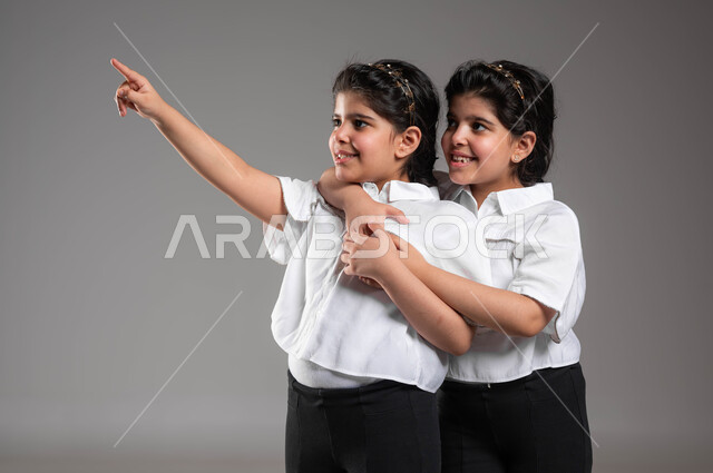 الشعور بالأمان والحب، العناق بين الشقيقات، مشاعر المحبة والود بين الاخوة، الاشارة الى شيء ما بعيد، بورتريه مقرب لفتاة عربية خليجية سعودية تشير بأصبعها للدلالة على شيء ما بجانب اختها التوأم، لفت الانتباه لشيء معين، خلفية رمادية