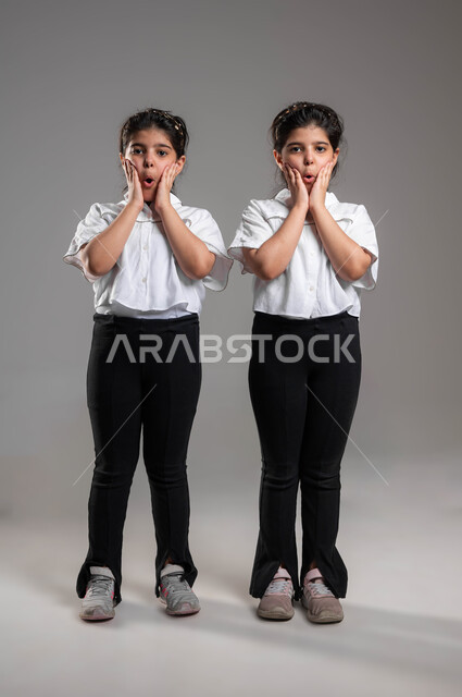 ايماءات التعجب والصدمة والذهول، الاستغراب من شيء ما، تلقي اخبار مفاجئة، بورتريه لأختين توأم عربيتين خليجيتين سعوديتين تنظران الى الكاميرا بتعابير الدهشة، ارتداء الزي الكاجوال والاهتمام بالمظهر الخارجي، صورة الجسم بالطول الكامل، خلفية رمادية