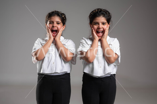 الاستغراب من شيء ما، الشعور بالتعجب والصدمة والذهول، تلقي اخبار مفرحة ومفاجئة، بورتريه مقرب لأختين توأم عربيتين خليجيتين سعوديتين تنظران الى الكاميرا بتعابير الدهشة والسرور، ارتداء الزي الكاجوال والاهتمام بالمظهر الخارجي، خلفية رمادية