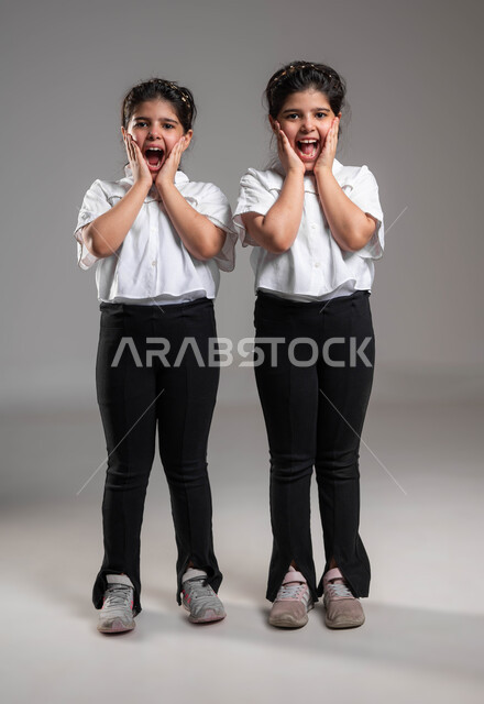 الشعور بالتعجب والصدمة والذهول، الاستغراب من شيء ما، تلقي اخبار مفرحة ومفاجئة، بورتريه لأختين توأم عربيتين خليجيتين سعوديتين تنظران الى الكاميرا بتعابير الدهشة والسرور، ارتداء الزي الكاجوال والاهتمام بالمظهر الخارجي، صورة الجسم بالطول الكامل، خلفية رمادية