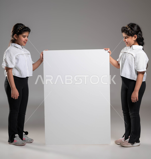 لوح اعلاني فارغ، استخدام لوحات توضيحية ووسائل تعليمية، إعلانات وعروض تسويقية، بورتريه لأختين توأم عربيتين خليجيتين سعوديتين مبتسمتين تقفان بجانب موك اب مستطيل كبير وتنظران اليه باهتمام، صورة الجسم بالطول الكامل، خلفية رمادية