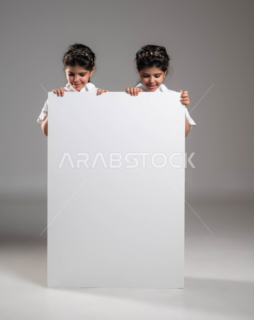 Use of explanatory panels and educational materials, advertisements and marketing presentations, an empty billboard, a portrait of two smiling Saudi Gulf Arab twin sisters standing behind a large rectangular mockup and looking at it with interest, a gray background.