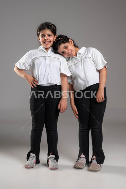 مفهوم مساندة ودعم الأخوات، تعزيز وتقوية الروابط العائلية، المحبة بين الشقيقات، معنى الصداقة والاخوة، بورتريه لبنت عربية خليجية سعودية تضع رأسها على كتف أختها التوأم وتمسك بيدها، صورة الجسم بالطول الكامل، خلفية رمادية
