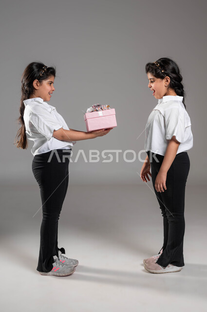 الترابط والمحبة بين الشقيقات، ايماءات السرور والمودة بالاحتفال في عيد الميلاد، تقوية وتعزيز الروابط العائلية، بورتريه  لبنت عربية خليجية سعودية تقوم بتقديم هدية لأختها التوأم، أعياد ومناسبات ومفاجآت سعيدة, صورة الجسم بالطول الكامل، خلفية رمادية