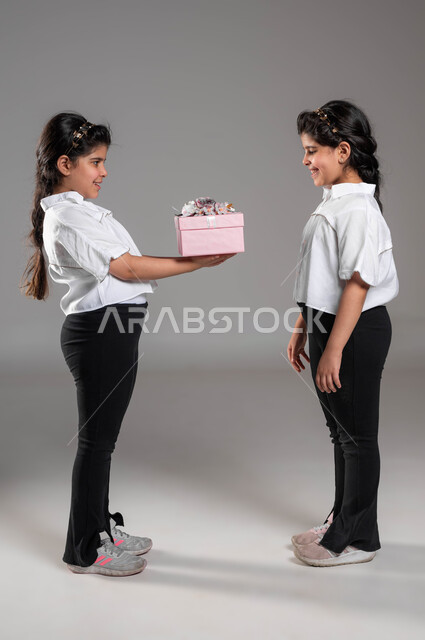 ايماءات السرور والمودة،  التعبير عن مشاعر المحبة بين الأخوة، تقوية وتعزيز الروابط العائلية، بورتريه لبنت عربية خليجية سعودية تقوم بتقديم هدية لأختها التوأم في يوم عيد ميلادها، أعياد ومناسبات ومفاجآت سعيدة, صورة الجسم بالطول الكامل، خلفية رمادية