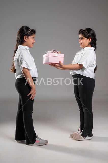 اظهار الدعم للأخت بالاحتفال بعيد ميلادها، مفهوم الترابط والمحبة بين الأخوة، تعزير الروابط العائلية وتقوية العلاقة بين الأشقاء، بورتريه لبنت عربية خليجية سعودية تقوم بتقديم هدية لأختها التوأم بمناسبة يوم ميلادها، صورة الجسم بالطول الكامل، خلفية رمادية