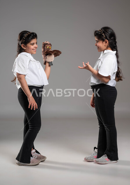 الاستمتاع بالأنشطة الترفيهية وقضاء أوقات مسلية، بورتريه لأختين توأم عربيتين خليجيتين سعوديتين مبتسمتين تقومان باللعب في دمية اليد القماشية، التدرب على العروض المسرحية، ايماءات وتعابير الاستفسار والتساؤل، صورة لكامل الجسم، خلفية رمادية
