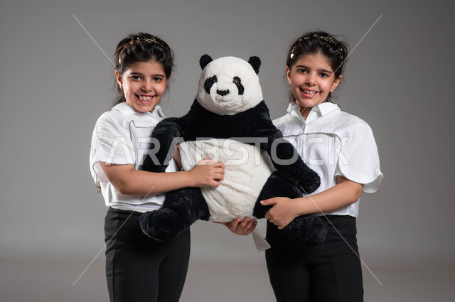 قضاء أوقات ممتعة ومسلية، بورتريه مقرب لأختين توأم عربيتين خليجيتين سعوديتين مبتسمتين تمسكان دمية دب الباندا القماشية تنظران الى الكاميرا، الاستمتاع بالأنشطة الترفيهية والألعاب، ايماءات وتعابير السعادة والسرور، خلفية رمادية