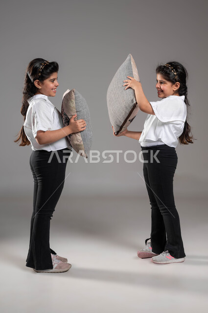 قضاء أوقات ممتعة ومسلية، بورتريه لتوأم بنات عربيات خليجيات سعوديات مبتسمات تقومان باللعب واللهو في الوسادات، ايماءات وتعابير السعادة والسرور، صورة الجسم بالطول الكامل، الاستمتاع بالأنشطة الترفيهية والألعاب، خلفية رمادية