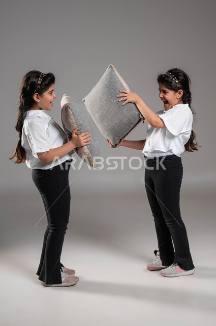 قضاء أوقات ممتعة ومسلية، بورتريه لتوأم بنات عربيات خليجيات سعوديات مبتسمات تقومان باللعب واللهو في الوسادات، ايماءات وتعابير السعادة والسرور، صورة الجسم بالطول الكامل، الاستمتاع بالأنشطة الترفيهية والألعاب، خلفية رمادية