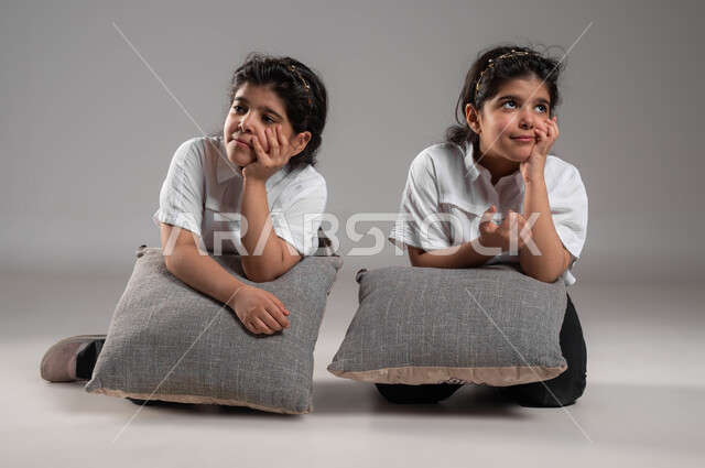 انتظار شيء ما، التفكير وشرود الذهن، الشعور والاحساس بالضجر والزهق، بورتريه لأختين توأم عربيتين خليجيتين سعوديتين تجلسان بملل وايماءات العبوس والانزعاج، خلفية رمادية