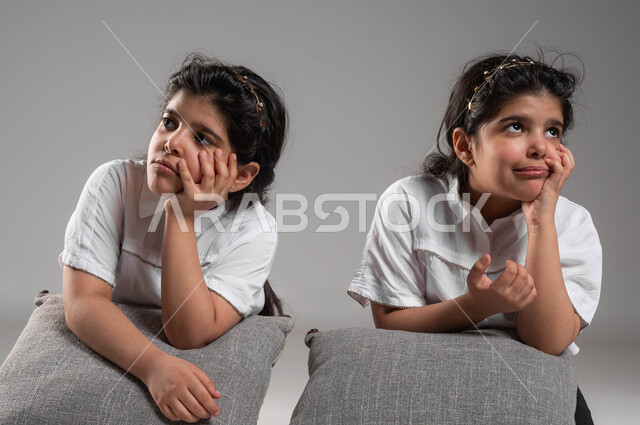 التفكير وشرود الذهن، الشعور والاحساس بالضجر والزهق، انتظار شيء ما، بورتريه لأختين توأم عربيتين خليجيتين سعوديتين تجلسان بملل وايماءات العبوس والانزعاج، خلفية رمادية