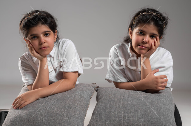 شرود الذهن والتفكير، الشعور والاحساس بالضجر والزهق، انتظار شيء ما، بورتريه لأختين توأم عربيتين خليجيتين سعوديتين تجلسان بملل وايماءات العبوس والانزعاج، خلفية رمادية