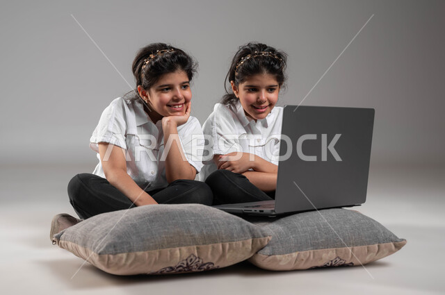 فعاليات ترفيهية الكترونية للاطفال، قضاء اوقات مسلية باللعب في الأجهزة التكنولوجية الحديثة، بورتريه لتوأم بنات عربيات خليجيات سعوديات مبتسمات يقومون بمشاهدة الأفلام والمقاطع المسلية  على الحاسوب المحمول، تعابير وايماءات التسلية والمتعة، خلفية رمادية