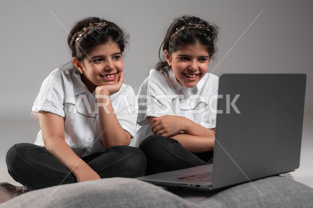 انشطة ترفيهية الكترونية للاطفال، قضاء اوقات مسلية باللعب في الأجهزة التكنولوجية الحديثة، بورتريه لتوأم بنات عربيات خليجيات سعوديات مبتسمات يقومون بمشاهدة الأفلام والمقاطع المسلية على الحاسوب المحمول، تعابير وايماءات التسلية والمتعة، خلفية رمادية