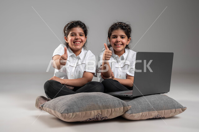 Feeling satisfied and privileged, watching movies and entertaining clips on the laptop, expressions of agreement and acceptance, portrait of twin Saudi Gulf Arab girls, smiling, raising a thumbs up as a sign of admiration, looking at the camera, using modern technological devices, gray background