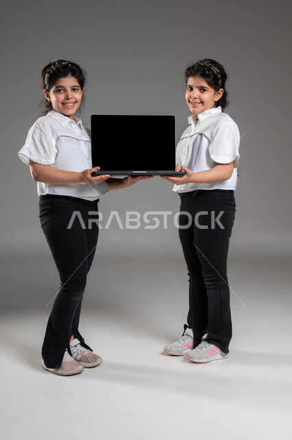 دور التقنيات الحديثة في تزويد الأجيال بالمعلومات المفيدة، استخدام الأجهزة التقنية المتطورة، بورتريه لأختين توأم عربيتين خليجيتين سعوديتين مبتسمتين تقومان بعرض حاسب محمول بشاشة سوداء فارغة، ايماءات السعادة والسرور، صورة الجسم بالطول الكامل، خلفية رمادية