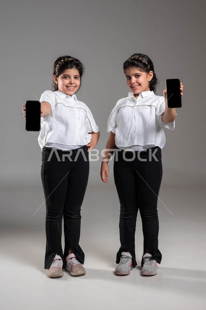 دور التقنيات الحديثة في تزويد الأجيال بالمعلومات المفيدة، استخدام الأجهزة التقنية المتطورة، بورتريه لأختين توأم عربيتين خليجيتين سعوديتين مبتسمتين تقومان بعرض هاتف محمول بشاشة سوداء فارغة، ايماءات السعادة والسرور، صورة الجسم بالطول الكامل، خلفية رمادية
