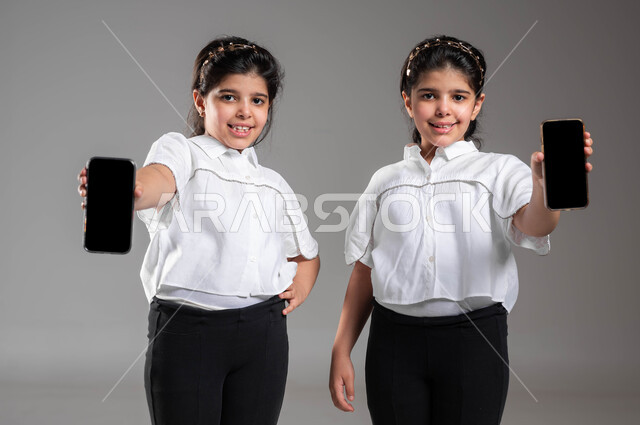 استخدام الأجهزة التقنية المتطورة، بورتريه مقرب لأختين توأم عربيتين خليجيتين سعوديتين مبتسمتين تقومان بعرض هاتف محمول بشاشة سوداء فارغة، دور التقنيات الحديثة في تزويد الأجيال بالمعلومات المفيدة، ايماءات السعادة والسرور، خلفية رمادية