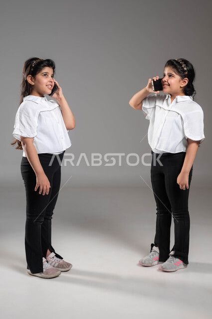 محادثات ودردشات مع الأصدقاء، ايماءات وتعابير الفرح والسرور بالتواصل مع الأقارب، بورتريه لأختين توأم عربيتين خليجيتين سعوديتين مبتسمتين تقومان بالتحدث باستخدام الجوال, إجراء مكالمه هاتفيه عبر الهاتف المحمول، صورة الجسم بالكامل، خلفية رمادية