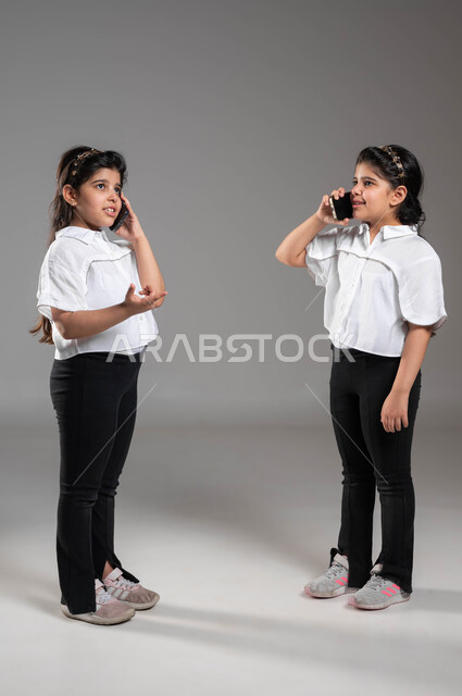 دردشه ومحادثات مع الأصدقاء, إجراء مكالمه هاتفيه عبر الهاتف المحمول، بورتريه لأختين توأم عربيتين خليجيتين سعوديتين مبتسمتين تقومان بالتحدث باستخدام الجوال، ايماءات وتعابير الفرح والسرور بالتواصل مع الأقارب، صورة الجسم بالكامل، خلفية رمادية