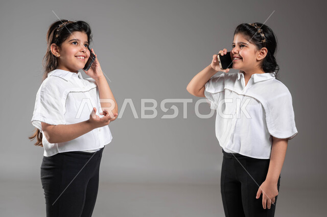 إجراء مكالمه هاتفيه عبر الهاتف المحمول، بورتريه مقرب لأختين توأم عربيتين خليجيتين سعوديتين مبتسمتين تقومان بالتحدث باستخدام الجوال، دردشه ومحادثات مع الأصدقاء, ايماءات وتعابير الفرح والسرور بالتواصل مع الأقارب، خلفية رمادية
