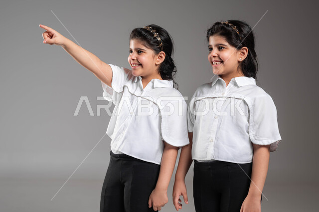 الفرح والسعادة لرؤية شيء بعيد، بورتريه مقرب لفتاة عربية خليجية سعودية مبتسمة تشير بأصبعها للدلالة على شيء ما بجانب اختها التوأم، لفت الانتباه لشيء معين، ايماءات وتعابير الاعجاب والسرور، خلفية رمادية