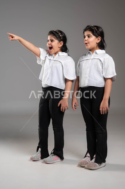 التعجب والدهشة لرؤية شيء بعيد، بورتريه لفتاة عربية خليجية سعودية تشير بأصبعها للدلالة على شيء ما بجانب اختها التوأم، لفت الانتباه لشيء معين، ايماءات وتعابير الصدمة والذهول، صورة الجسم بالطول الكامل، خلفية رمادية