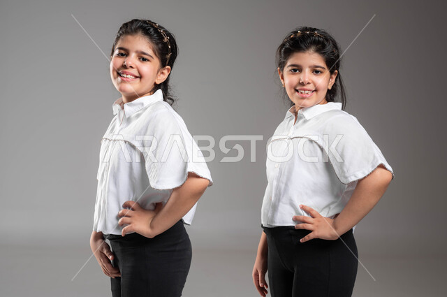 ارتداء الزي الكاجوال والاهتمام بالمظهر الخارجي، مفهوم التوأم الحقيقي المتطابق، بورتريه مقرب لتوأم بنات عربيات خليجيات سعوديات مبتسمات، الوقوف باستقامة مع وضع اليد على الخصر والنظر الى الكاميرا، ايماءات وتعابير الثقة بالنفس، خلفية رمادية