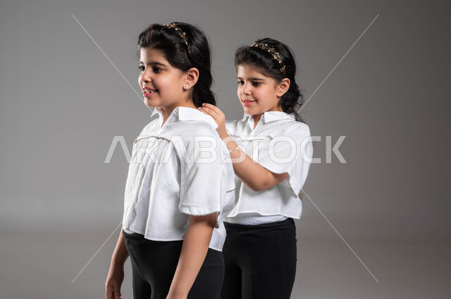 Paying attention and care to the external appearance, getting ready and prepared to go somewhere, the concept of a true identical twin, a close-up portrait of a Saudi Arabian Gulf girl arranging and styling her sister’s hair, bonding and brotherly cooperation, gestures and expressions of pleasure and joy, a gray background.