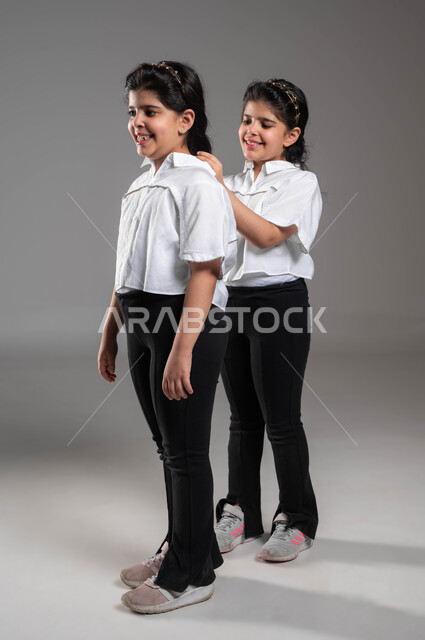 Getting ready and preparing to go somewhere, the concept of a real identical twin, a portrait of a Saudi Arabian Gulf girl arranging and styling her sister’s hair, bonding and brotherly cooperation, attention and care for the external appearance, gestures and expressions of pleasure and joy, a full-body portrait, gray background