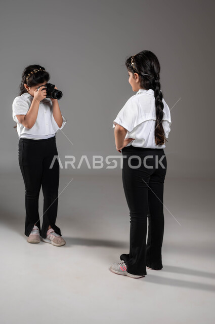 توثيق اللحظات السعيدة، بورتريه لأختين توأم عربيتين خليجيتين سعوديتين مبتسمتين تقومان بجلسة تصوير باستخدام الكاميرا، الاستمتاع بالتقاط صور تذكارية، تنمية فنون ومواهب التصوير الفوتوغرافي، اختيار المهنة المستقبلية، صورة الجسم بالطول الكامل، خلفية رمادية