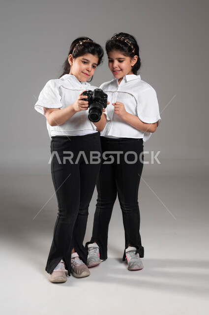 تنمية فنون ومواهب التصوير الفوتوغرافي، بورتريه لأختين توأم عربيتين خليجيتين سعوديتين مبتسمتين تقومان بمشاهدة الصور عبر الكاميرا الرقمية، الاستمتاع بالتقاط صور تذكارية، توثيق اللحظات السعيدة، اختيار المهنة المستقبلية، صورة الجسم بالطول الكامل، خلفية رمادية