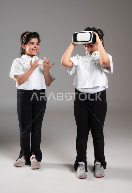 قضاء أوقات ممتعة ومسلية بالألعاب الافتراضية، بورتريه لفتاة عربية خليجية سعودية ترتدي نظارة في ار التقنية بجانب اختها التوأم، ايماءات وتعابير الاستفسار والفضول حول رؤية شيء ما، الاستمتاع بمشاهدة أفلام الواقع الافتراضي، صورة لكامل الجسم، خلفية رمادية
