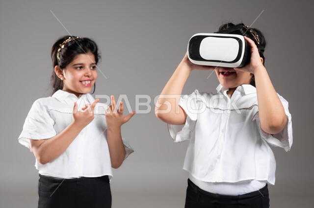 Enjoying watching virtual reality movies, spending fun and entertaining times playing virtual games, a close-up portrait of a Saudi Gulf Arab girl wearing VR technology glasses next to her twin sister, gestures and expressions of inquiry and curiosity about seeing something, gray background