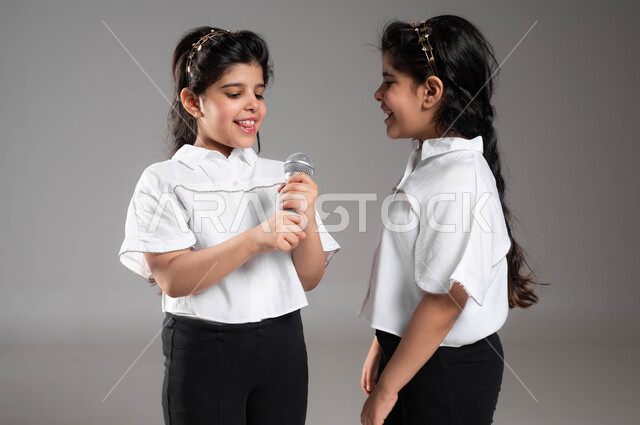 ايماءات وتعابير الاندماج والسرور، قضاء اوقات ممتعة ومسلية برفقة الاخوة, اكتشاف موهبة الغناء منذ الصغر, بورتريه مقرب لفتاة عربية سعودية خليجية تحمل بيدها الميكروفون تقوم بالغناء بجانب اختها التوأم، تقديم العروض الفنية المسرحية, خلفية رمادية