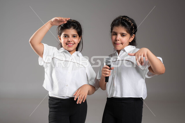 Spending fun and entertaining times with brothers, discovering the talent for singing since childhood, a close-up portrait of a Saudi Arabian Gulf girl holding a microphone in her hand, singing alongside her twin sister, gestures and expressions of integration and enthusiasm, presenting theatrical artistic performances, gray background