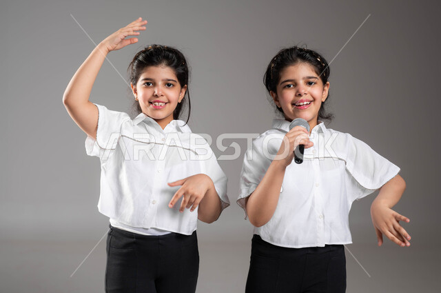 اكتشاف موهبة الغناء منذ الصغر, بورتريه مقرب لفتاة عربية سعودية خليجية تحمل بيدها الميكروفون تقوم بالغناء بجانب اختها التوأم، قضاء اوقات ممتعة ومسلية برفقة الاخوة, ايماءات وتعابير الاندماج والحماس، تقديم العروض الفنية المسرحية, خلفية رمادية