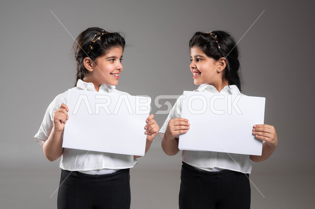 لوحة بيضاء مربعة فارغة، استخدام لوحات توضيحية تعريفية، إعلانات ودعايات وعروض تسويقية، بورتريه مقرب لأختين توأم عربيتين خليجيتين سعوديتين مبتسمتين تحملان موك اب مربع أبيض تنظران الى بعضهما بإيماءت السعادة و الفرح، خلفية رمادية