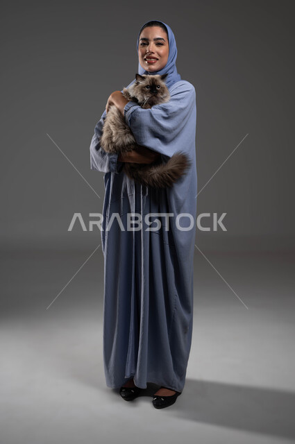 الاحساس بالسعادة أثناء قضاء وقت ممتع في اللعب مع القط، الرفق بالحيوان والعطف عليه، الأثر الايجابي لاقتناء الحيوانات المنزلية وتربيتها، بورتريه لشابة عربية خليجية سعودية مبتسمة ترتدي الحجاب والعباءة تقوم بحمل قطة رمادية أليفة، صورة لكامل الجسم، خلفية رمادية