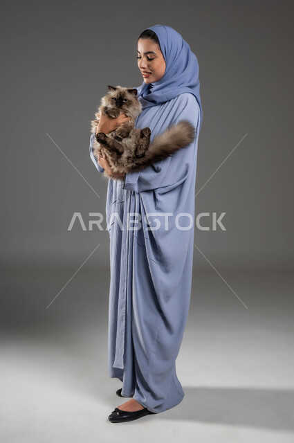 الاحساس بالسعادة أثناء قضاء وقت ممتع في اللعب مع القط، الرفق بالحيوان والعطف عليه، الأثر الايجابي لاقتناء الحيوانات المنزلية وتربيتها، بورتريه لشابة عربية خليجية سعودية مبتسمة ترتدي الحجاب والعباءة تقوم بحمل قطة رمادية أليفة، صورة لكامل الجسم، خلفية رمادية