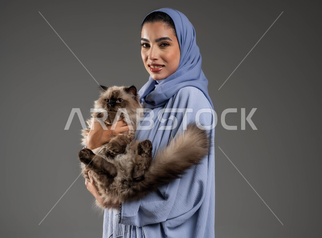مفهوم الرفق بالحيوان والعطف عليه، الأثر الايجابي لاقتناء الحيوانات المنزلية وتربيتها، بورتريه مقرب لإمرأة عربية خليجية سعودية مبتسمة ترتدي الحجاب والعباءة تقوم بحمل قطة رمادية أليفة، الشعور بالسعادة أثناء قضاء وقت ممتع في اللعب مع القط، خلفية رمادية