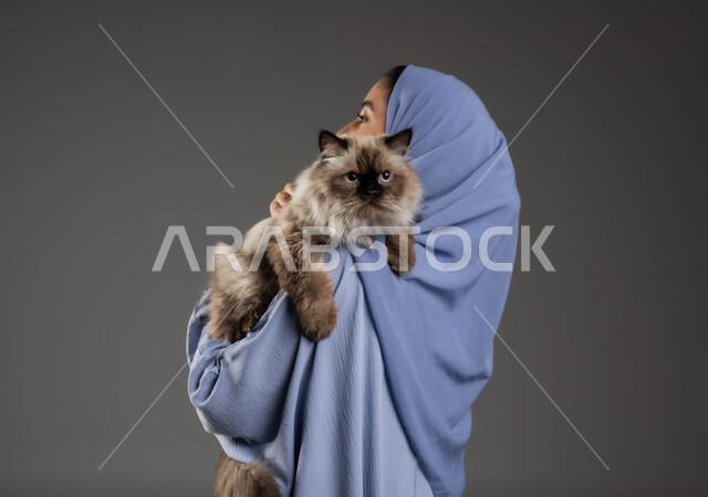 الشعور بالسعادة أثناء قضاء وقت ممتع في اللعب مع القط، بورتريه مقرب من الخلف لشابة عربية خليجية سعودية ترتدي الحجاب والعباءة تقوم بحمل قطة رمادية أليفة، مفهوم الرفق بالحيوان والعطف عليه، الأثر الايجابي لاقتناء الحيوانات المنزلية وتربيتها، خلفية رمادية
